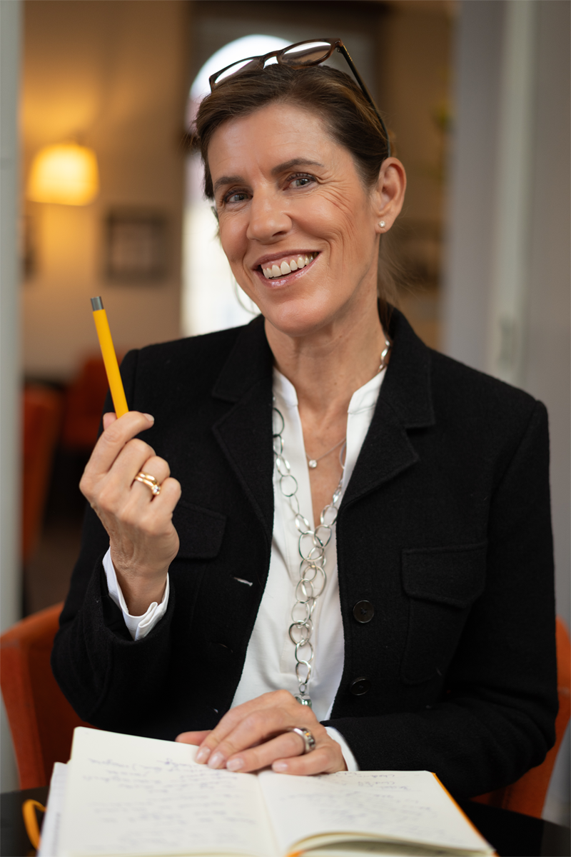 Maja Schneider sitzt mit Notizbuch am Tisch, hält einen Stift in der Hand und bereitet Inhalte für ein strukturiertes Kommunikationstraining vor.