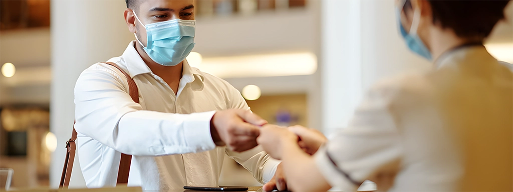 Hotelgast mit Maske überreicht seine Karte beim Check-in an die Rezeptionistin und erlebt einen strukturierten Empfang im Rahmen eines professionellen Front Office Checks.
